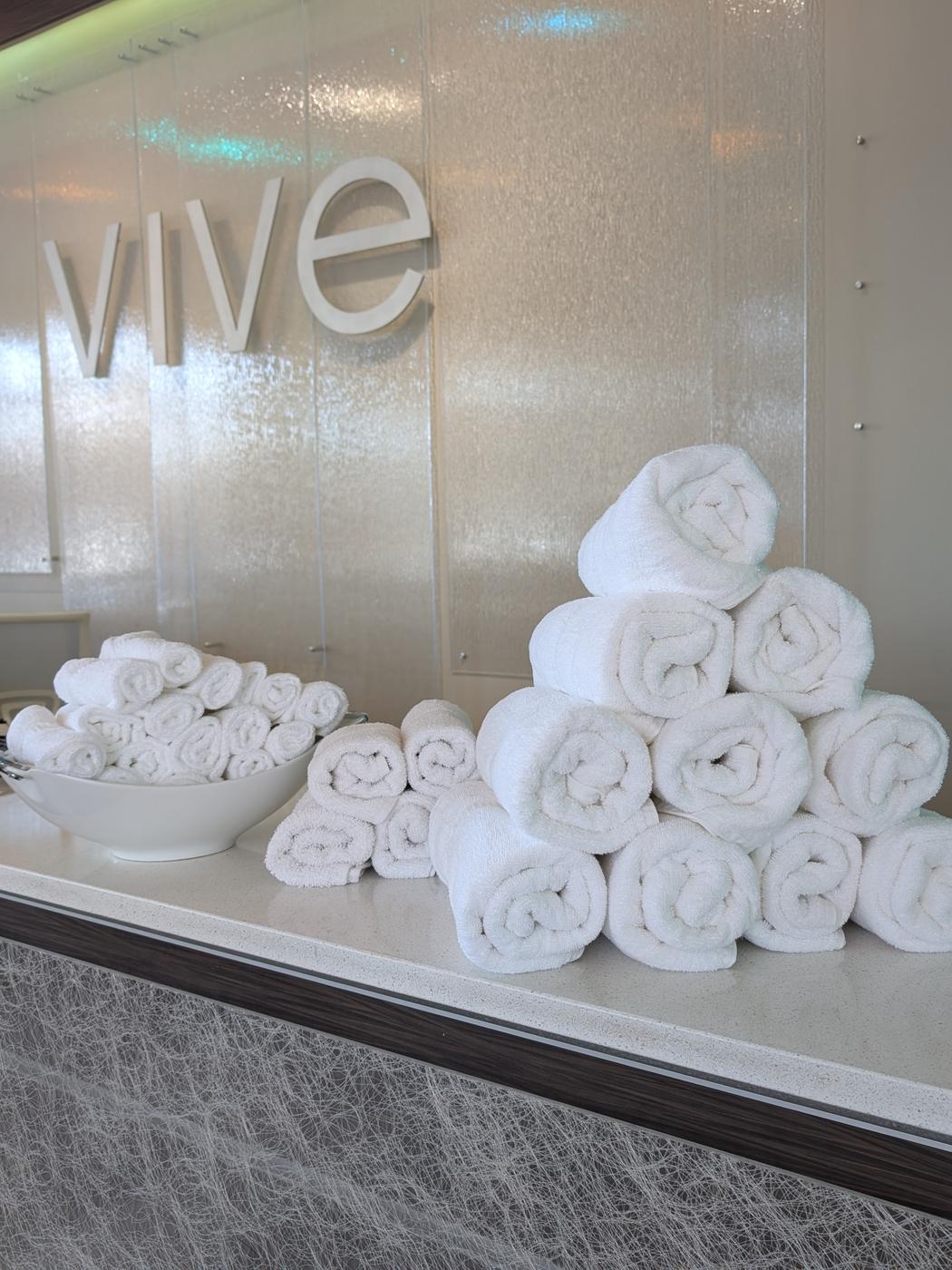 Rolled white towels stacked beneath the VIVE logo on a frosted glass wall